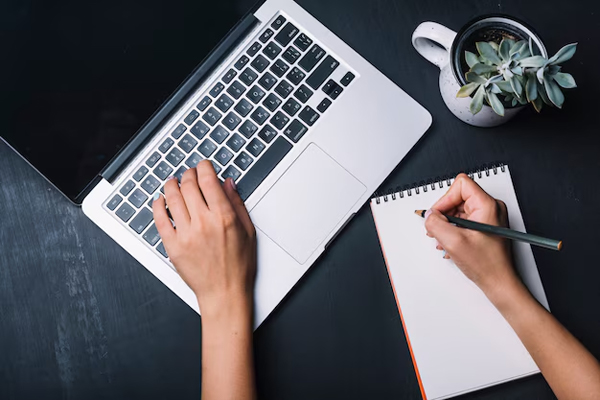 Top view of hands typing on laptop while writing notes in notebook with succulent plant on dark desk